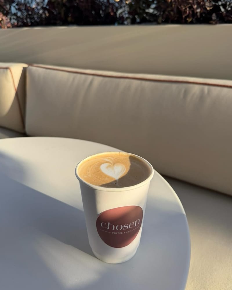 A takeaway cappuccino with heart latte art in a Chosen paper cup on a white table in the evening sun.