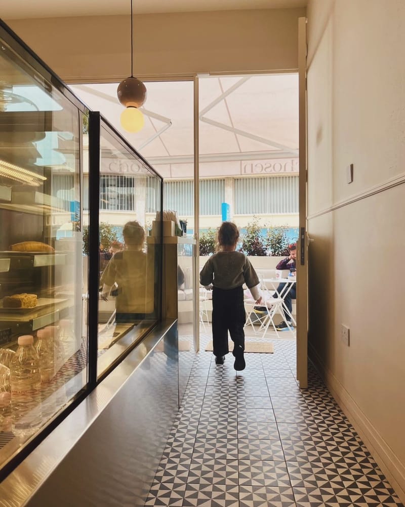 Looking down the shop past the pastry counter towards the bright street entrance, where a child walks out onto the terrace.