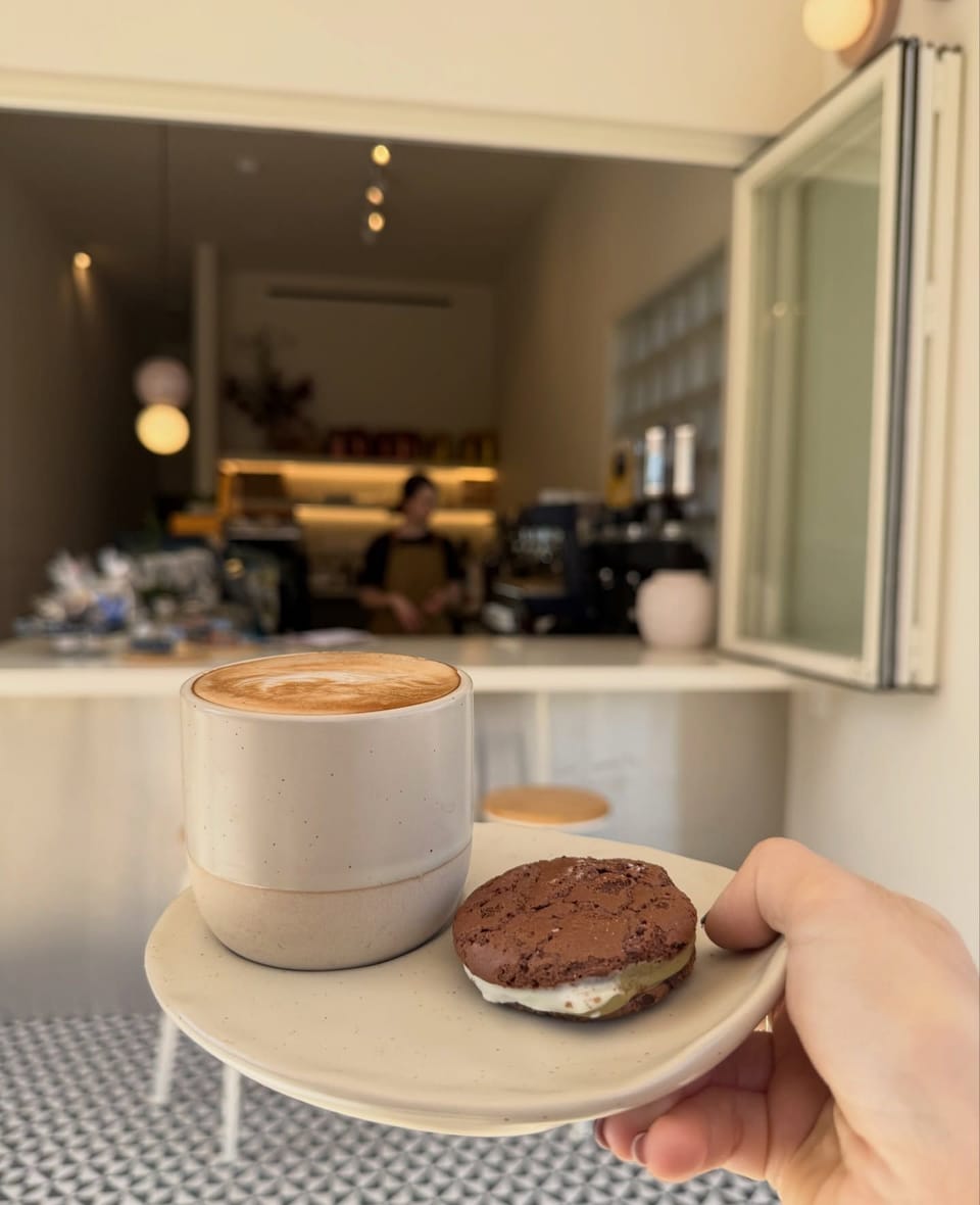 A flat white in a speckled cream cup with a chocolate cookie, held up in front of the open window of Chosen Coffee Shop, with the barista and counter behind.