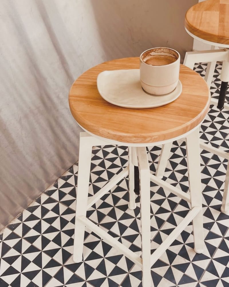 A cappuccino in a cream cup on a round wooden stool, standing on the shop's black and white geometric tiles.